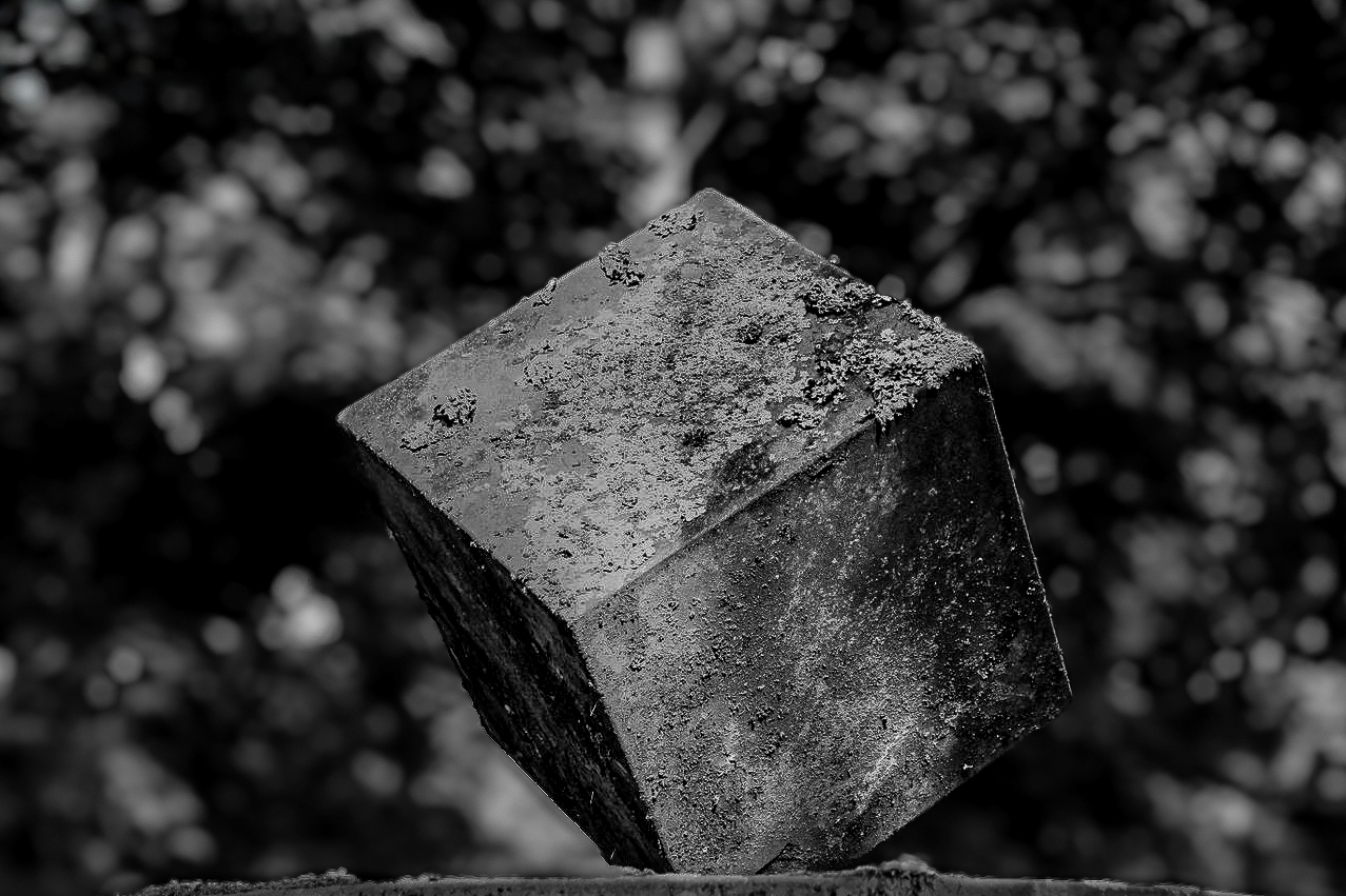 Schwarzweiß-Foto eines verwitterten, schräg stehenden Betonwürfels, der vor unscharfem, bokehartigem Hintergrund schwebend wirkt.