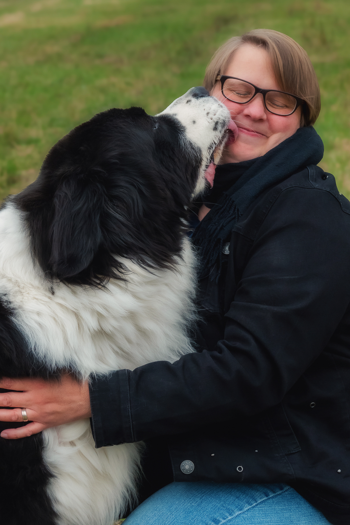 Hund gibt einer lachenden Frau einen liebevollen „Kuss“, inniger Moment zwischen Mensch und Tier auf einer Wiese, natürliche Farben und weiches Licht