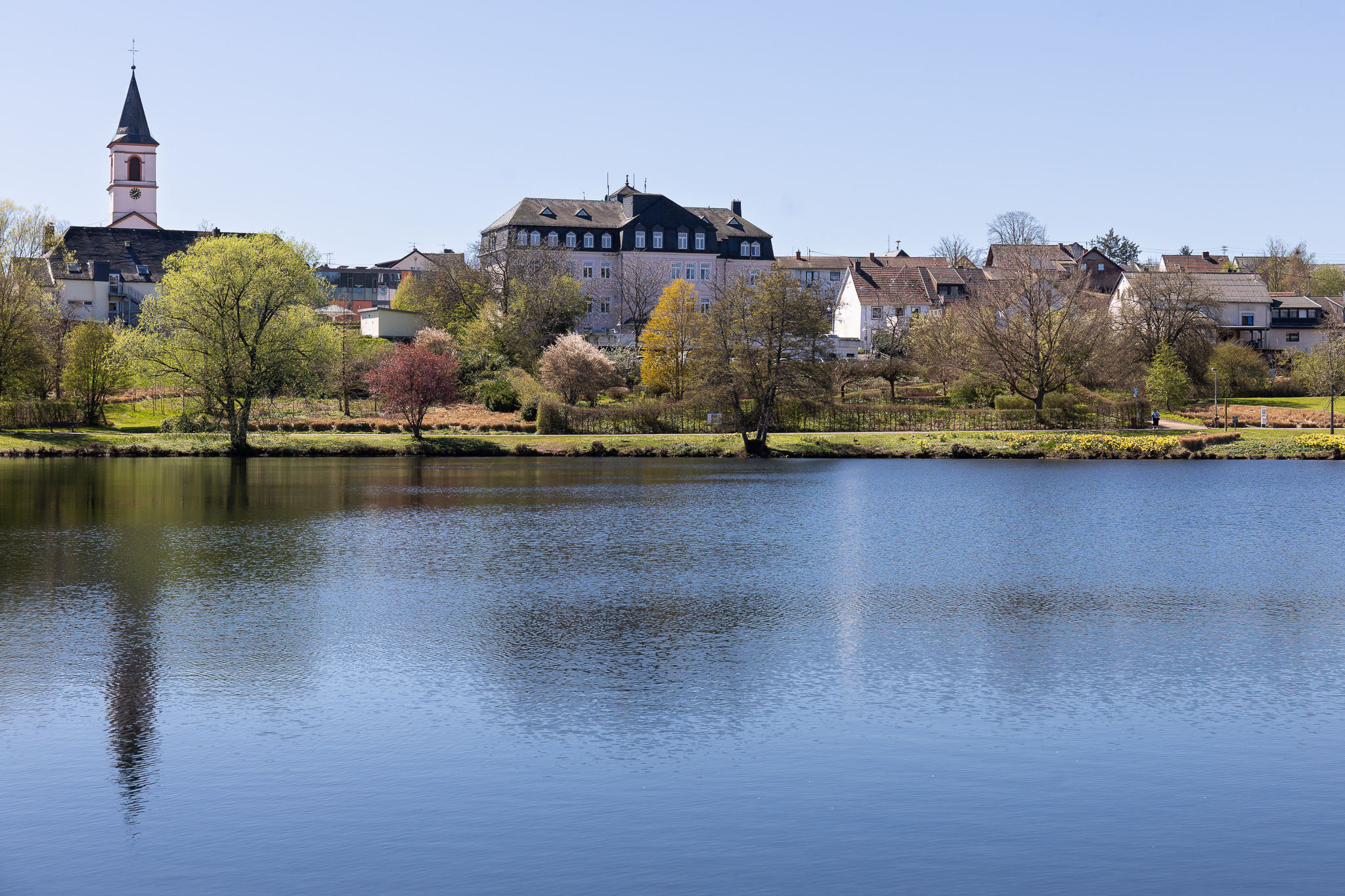 Blick über den Kurpark Weiskirchen mit Seeufer, Holzstruktur und Bäumen bei klarem Wetter